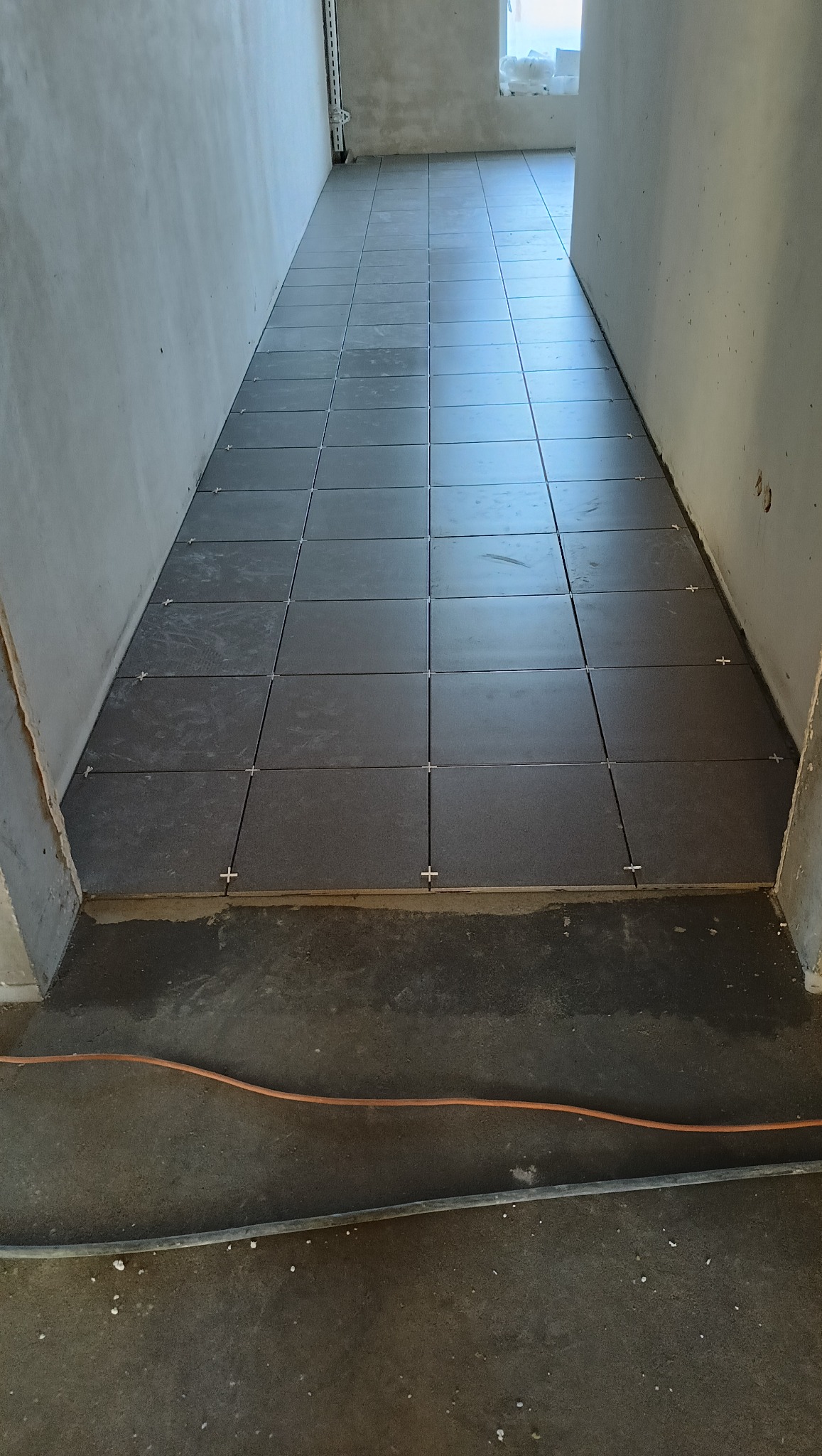 Układanie ciemnych, kwadratowych płytek podłogowych z krzyżykami dystansowymi w korytarzu. Widoczne surowe ściany i przewody. Etap prac wykończeniowych.