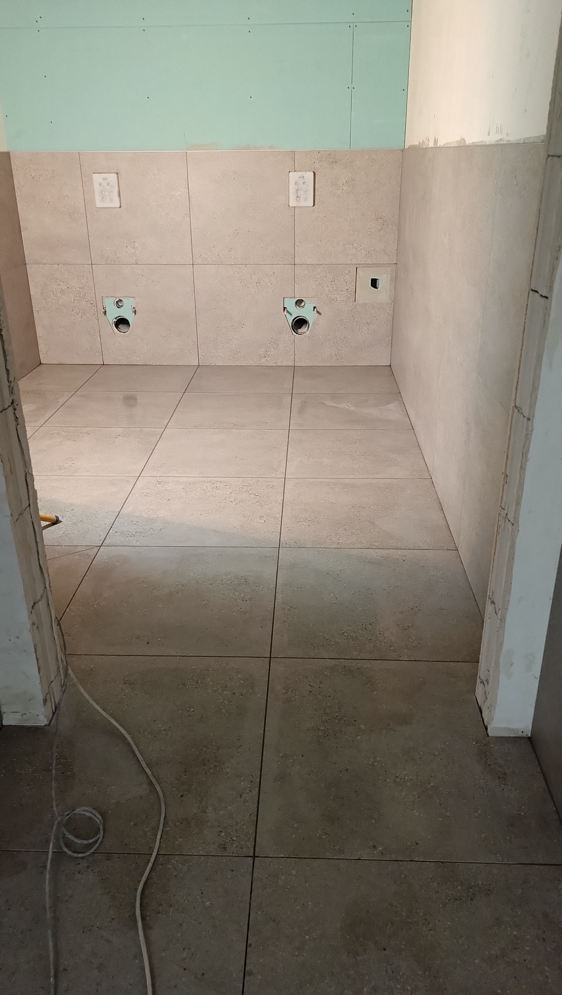 Wykończona beżowa łazienka z płytkami na podłodze i ścianach. Widoczne instalacje wodne i fragment ściany z płyt karton-gips. Przewód elektryczny na podłodze.