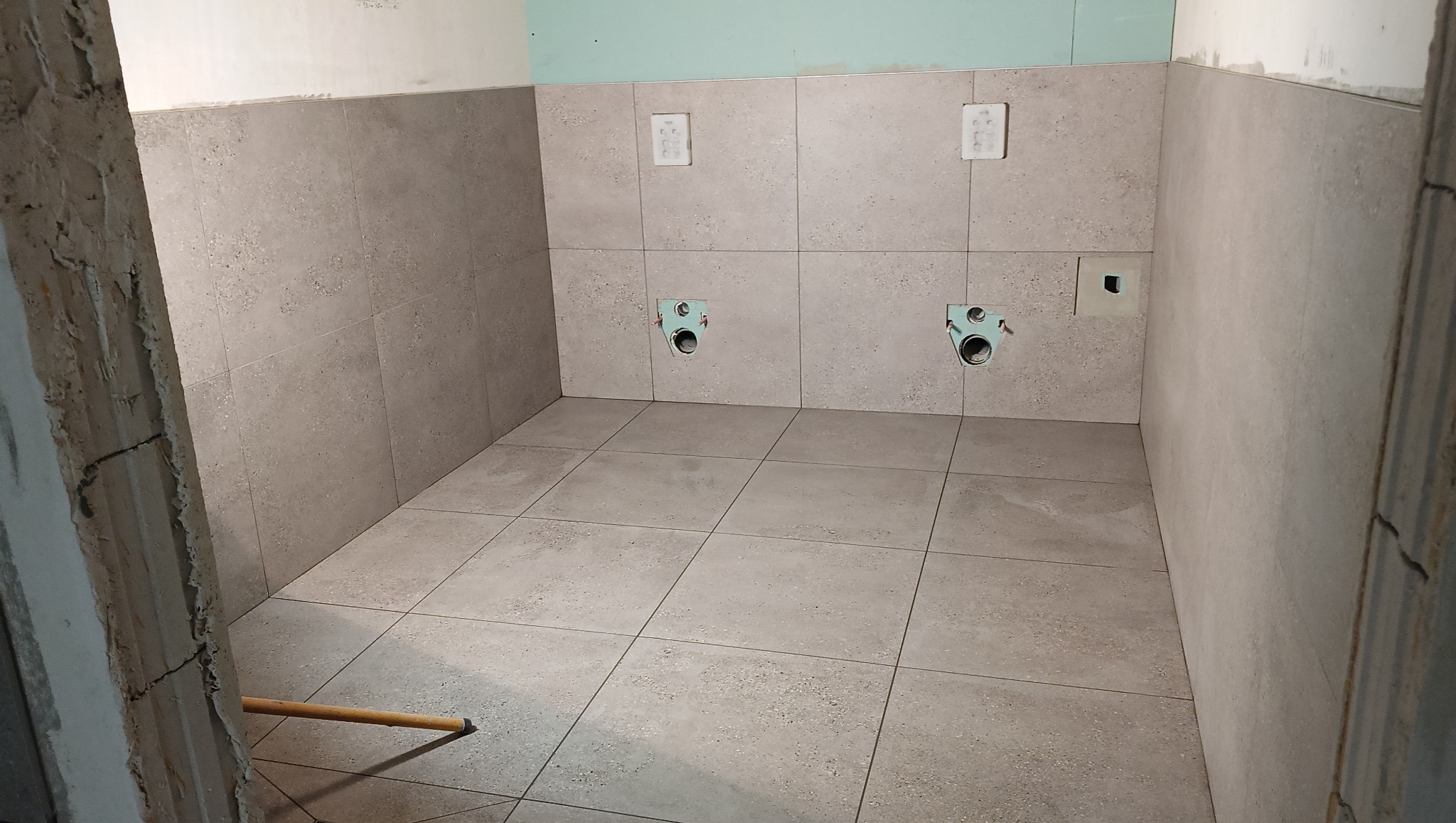 Wykończona łazienka z szarymi płytkami na ścianach i podłodze, widoczne przyłącza do toalety. Fragment ściany w stanie surowym.