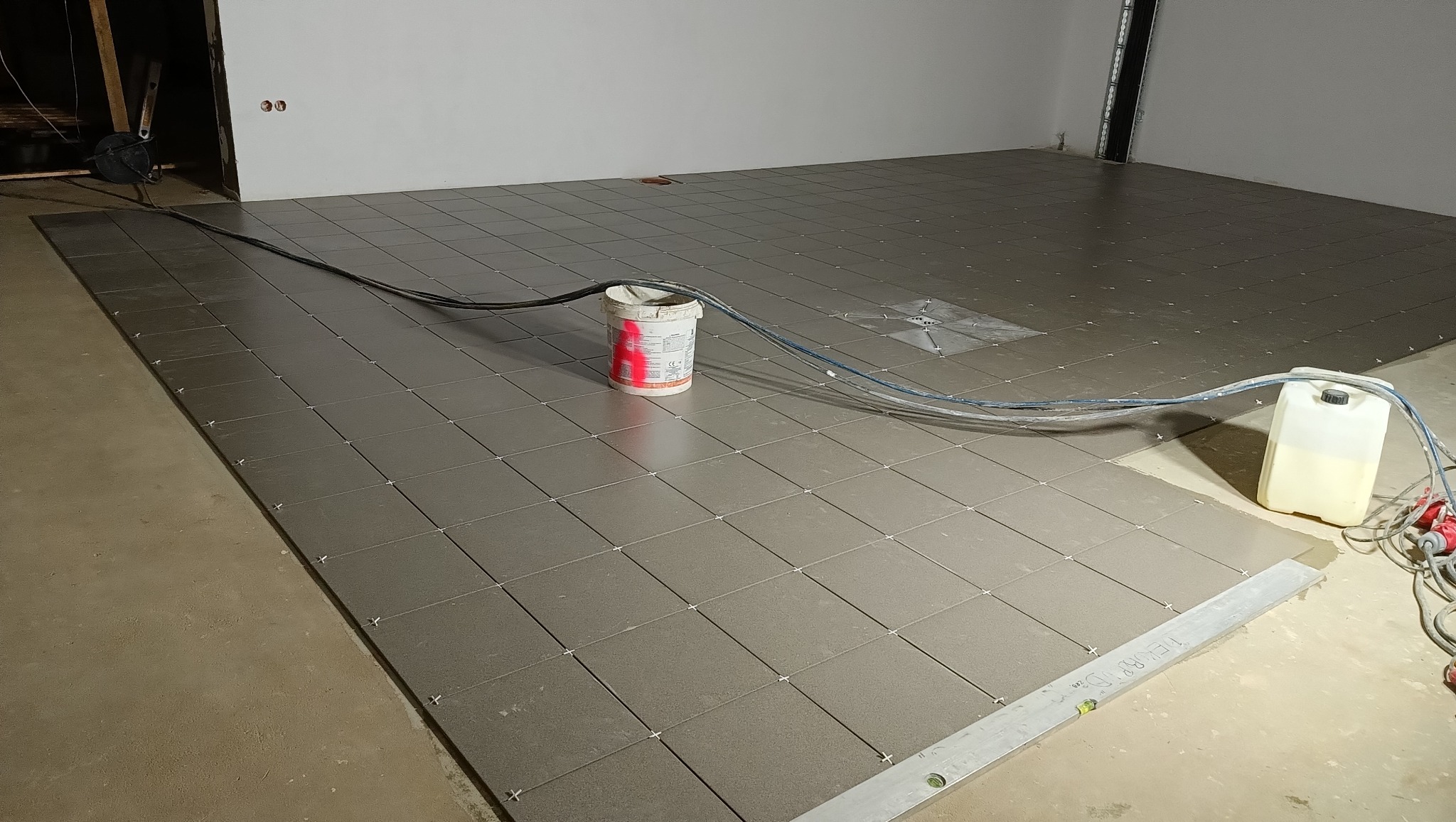 Układanie płytek podłogowych w trakcie prac. Widoczne krzyżyki dystansowe, poziomica i pojemniki z materiałami budowlanymi. Szare płytki na betonowej wylewce.