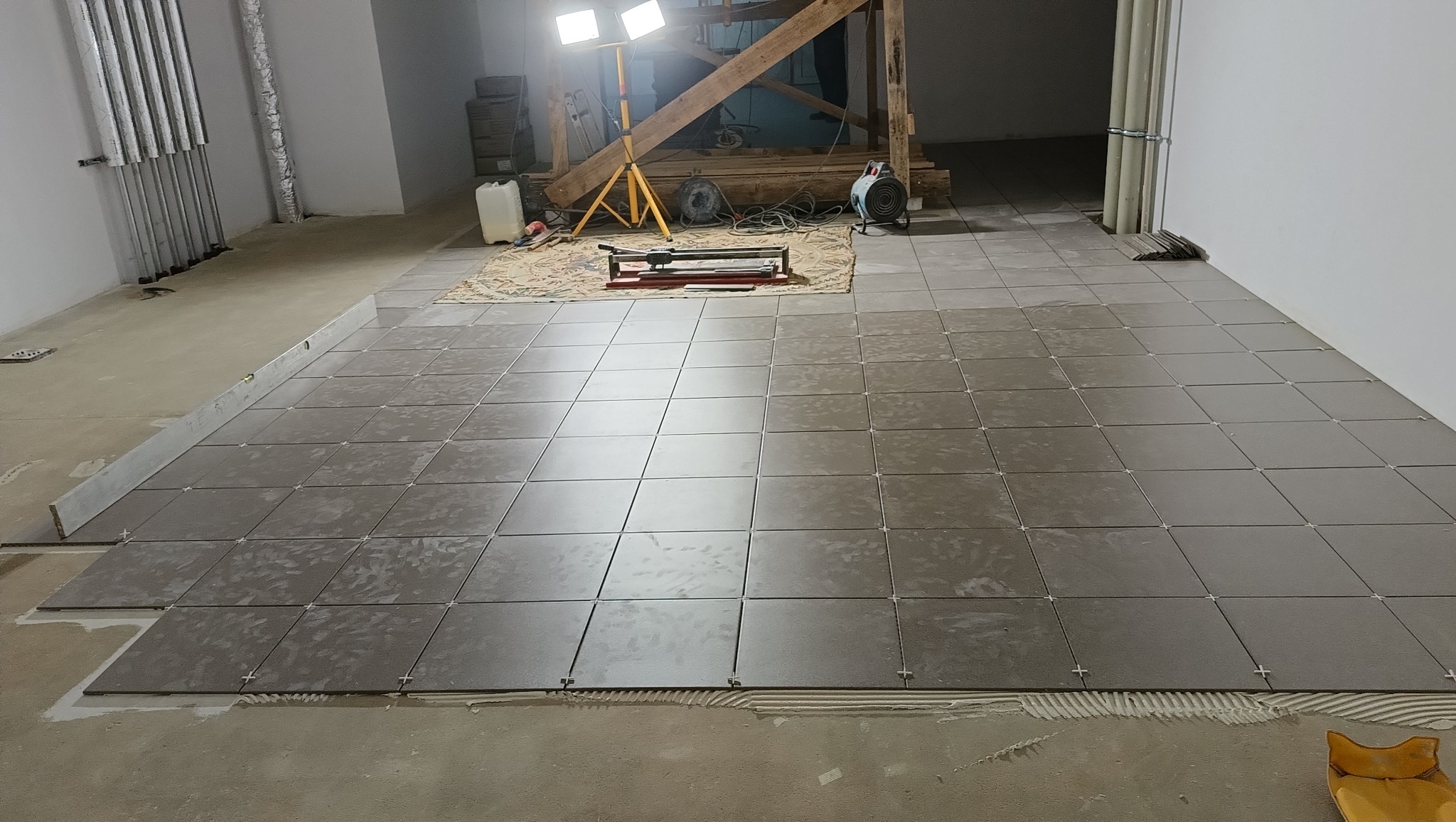 Układanie kwadratowych, matowych płytek podłogowych w szarym odcieniu. Widoczne krzyżyki dystansowe, narzędzia glazurnicze i oświetlenie budowlane w tle.
