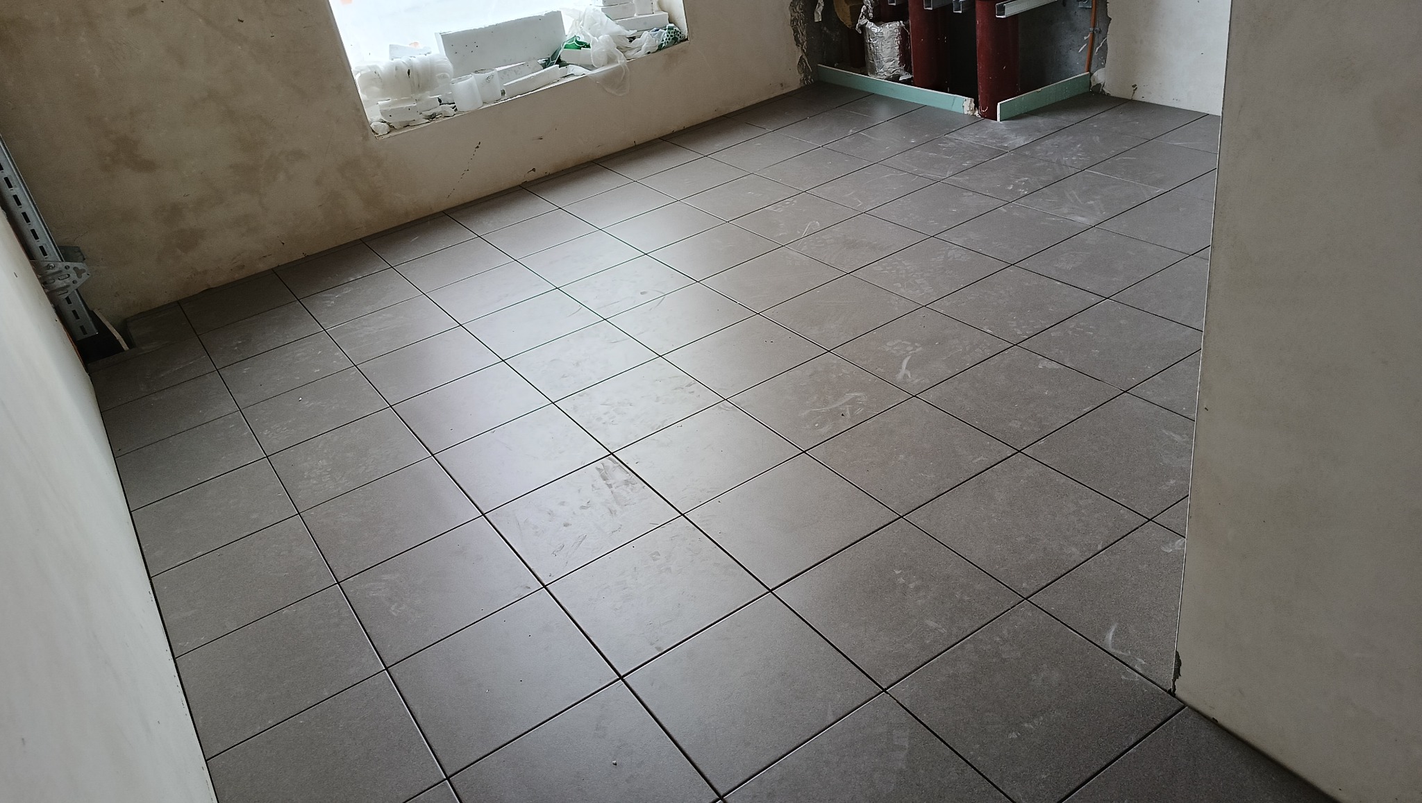 Ułożone szare płytki podłogowe w trakcie remontu łazienki, widoczne fragmenty ścian w surowym stanie i okno z materiałami budowlanymi.