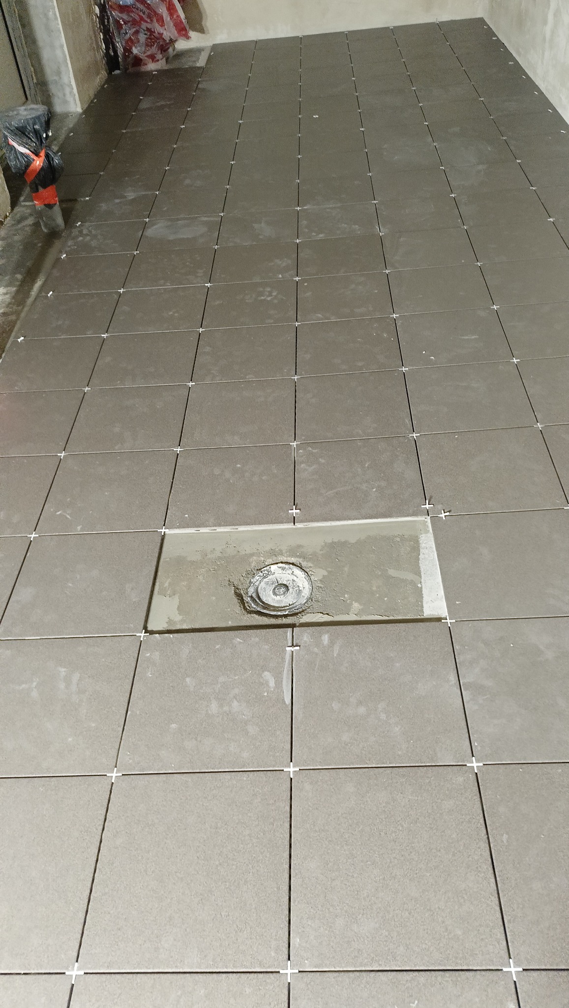 Układanie kwadratowych, szarych płytek podłogowych z białymi krzyżykami dystansowymi. W tle widoczny niezabudowany odpływ podłogowy.