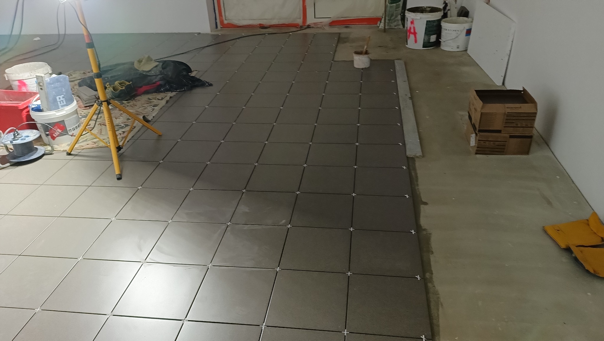 Układanie ciemnych płytek podłogowych z krzyżykami dystansowymi, widoczna lampa robocza, wiaderka i kartonowe pudełko w tle. Etap prac wykończeniowych.