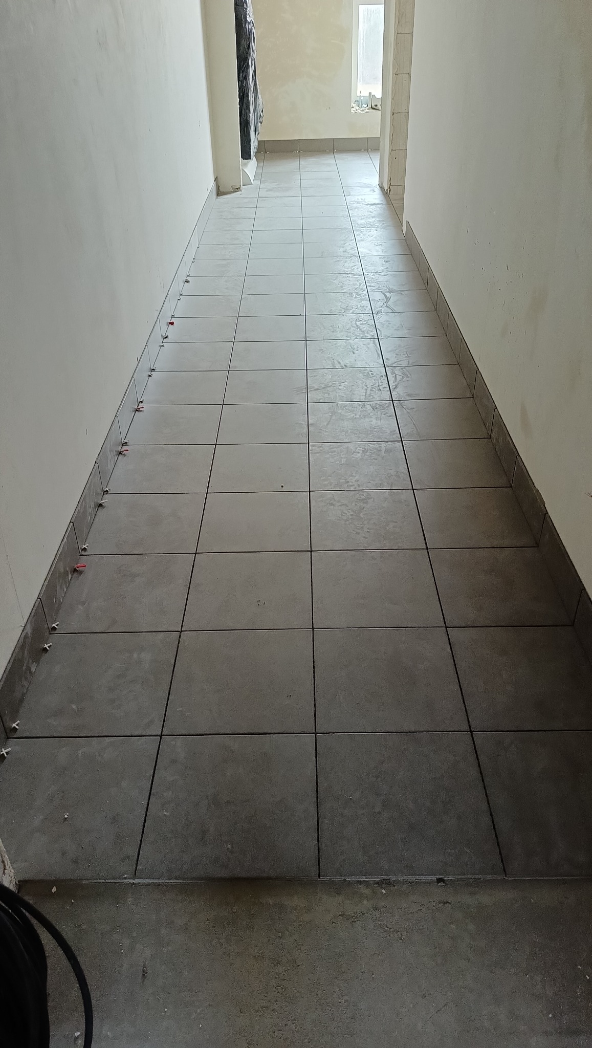 Korytarz w trakcie układania szarych płytek podłogowych. Widoczne krzyżyki dystansowe, jasne ściany i fragment niezakończonej podłogi.