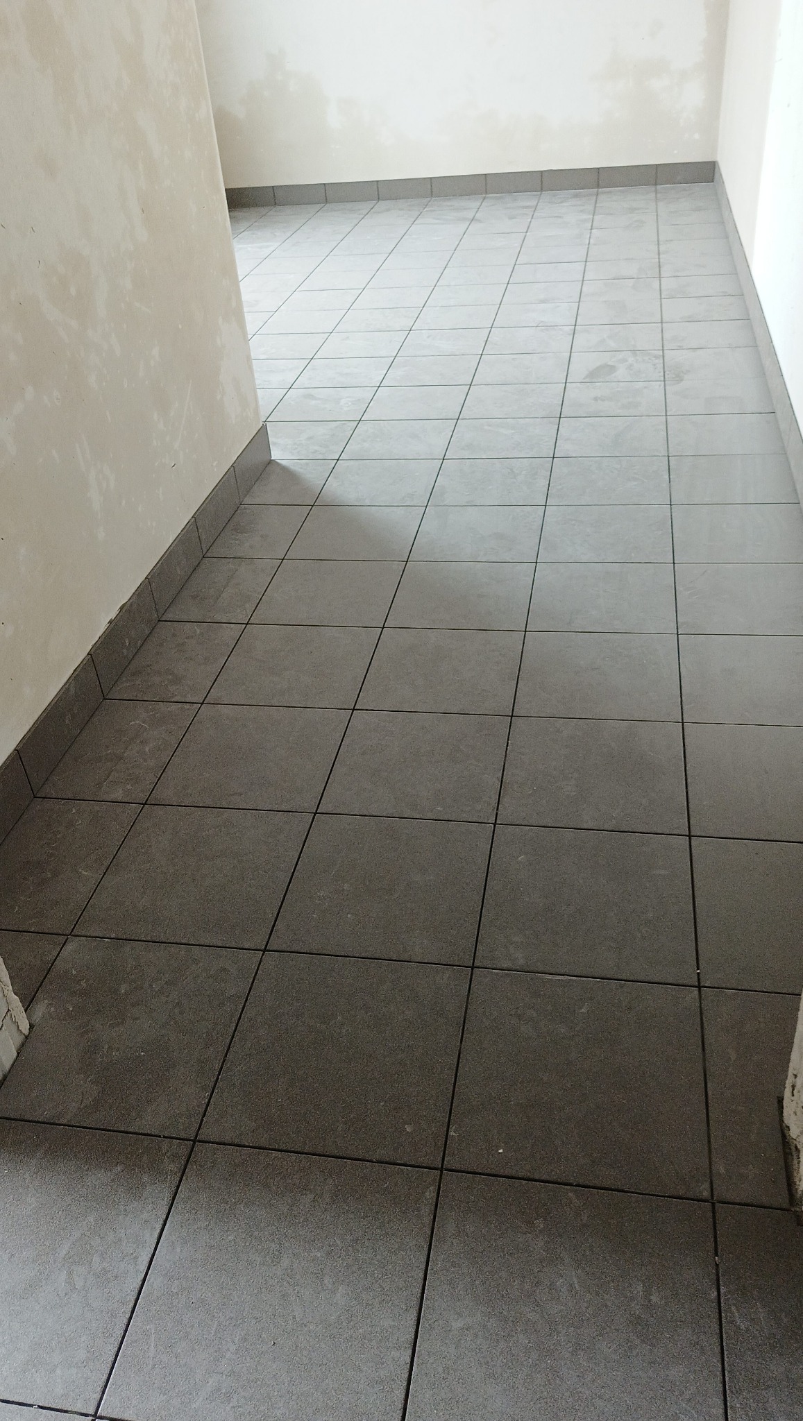 Ułożona, szara, kwadratowa terakota podłogowa z fugami w korytarzu; widoczne przypodłogowe płytki. Ściany w stanie surowym, przygotowane do dalszych prac.