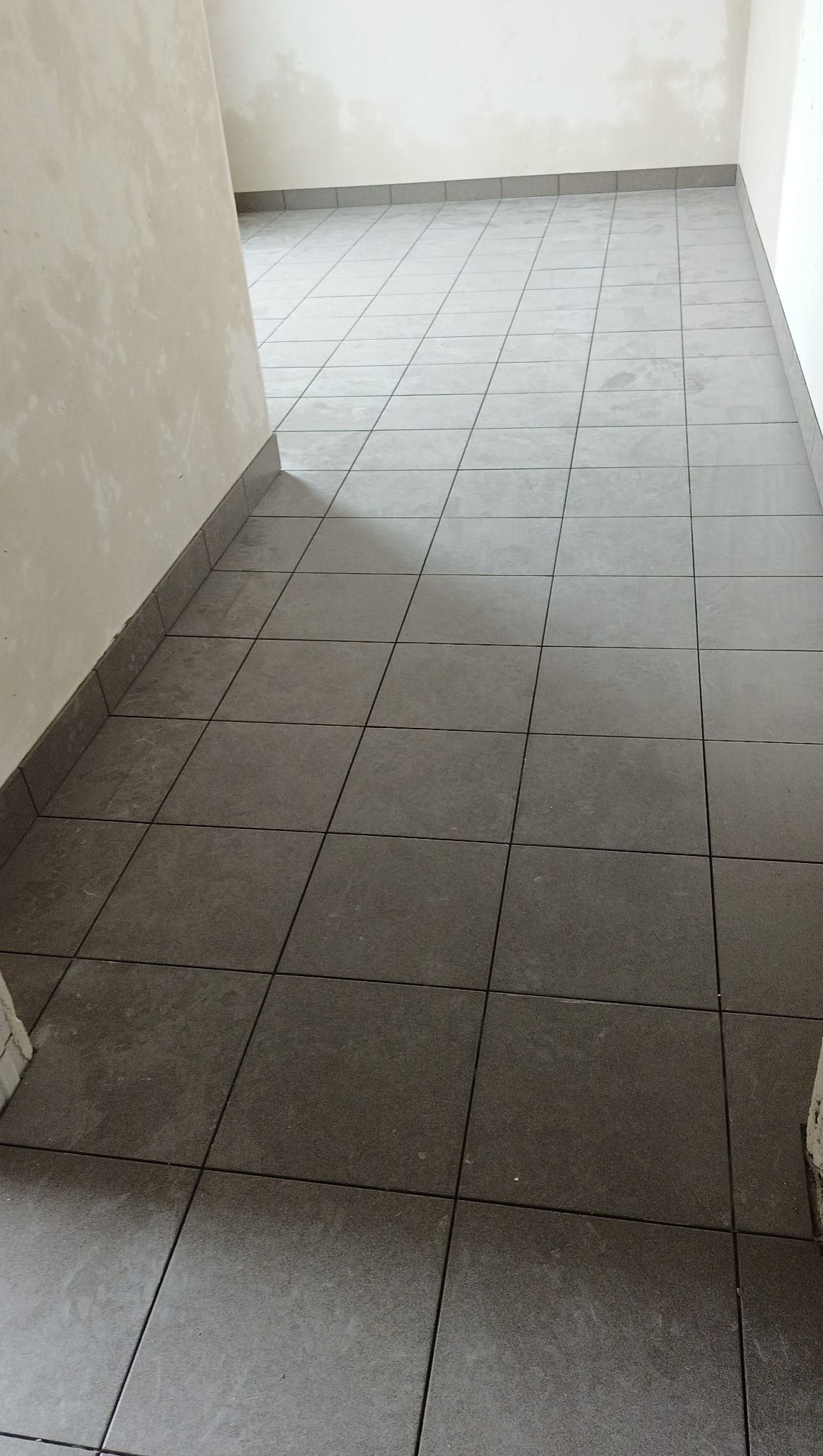 Ułożona podłoga z szarych, kwadratowych płytek w korytarzu. Widoczne niedokończone ściany w jasnym kolorze. Minimalistyczny styl wykończenia.