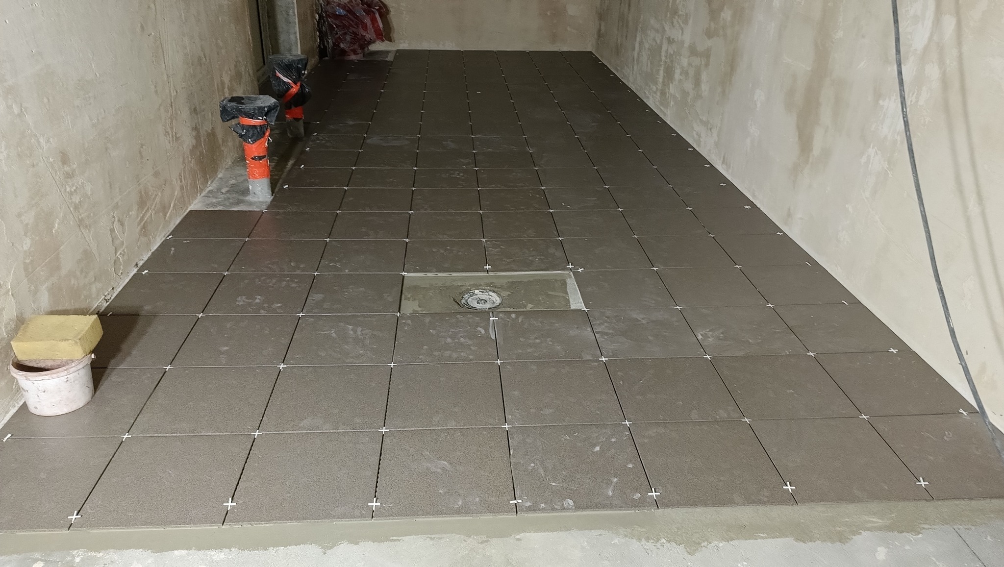 Układanie ciemnych, kwadratowych płytek na podłodze, z krzyżykami dystansowymi, widoczny odpływ liniowy, ściany surowe.