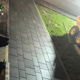 Ułożona szara kostka brukowa wzdłuż budynku z małym ogrodem, widoczny fragment maszyny budowlanej w tle.