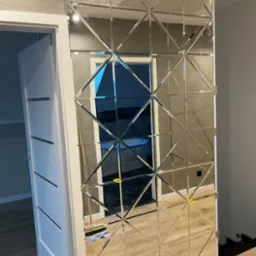 Ściana z dekoracyjnych luster w kształcie rombów, odbijająca wnętrze pokoju z widoczną białą ramą drzwi i fragmentem podłogi, zamontowana przy szarej ścianie.