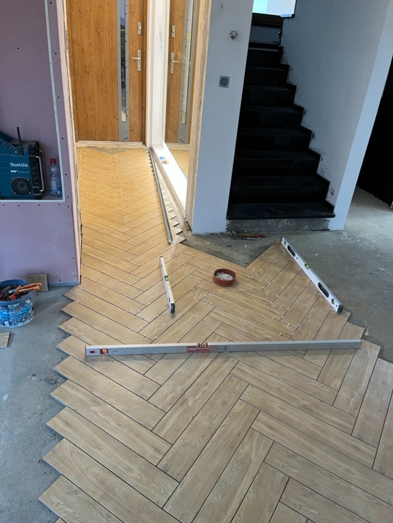 Układanie parkietu w jodełkę z płytek imitujących drewno w korytarzu, widoczne poziomice i narzędzia, w tle schody i drzwi.