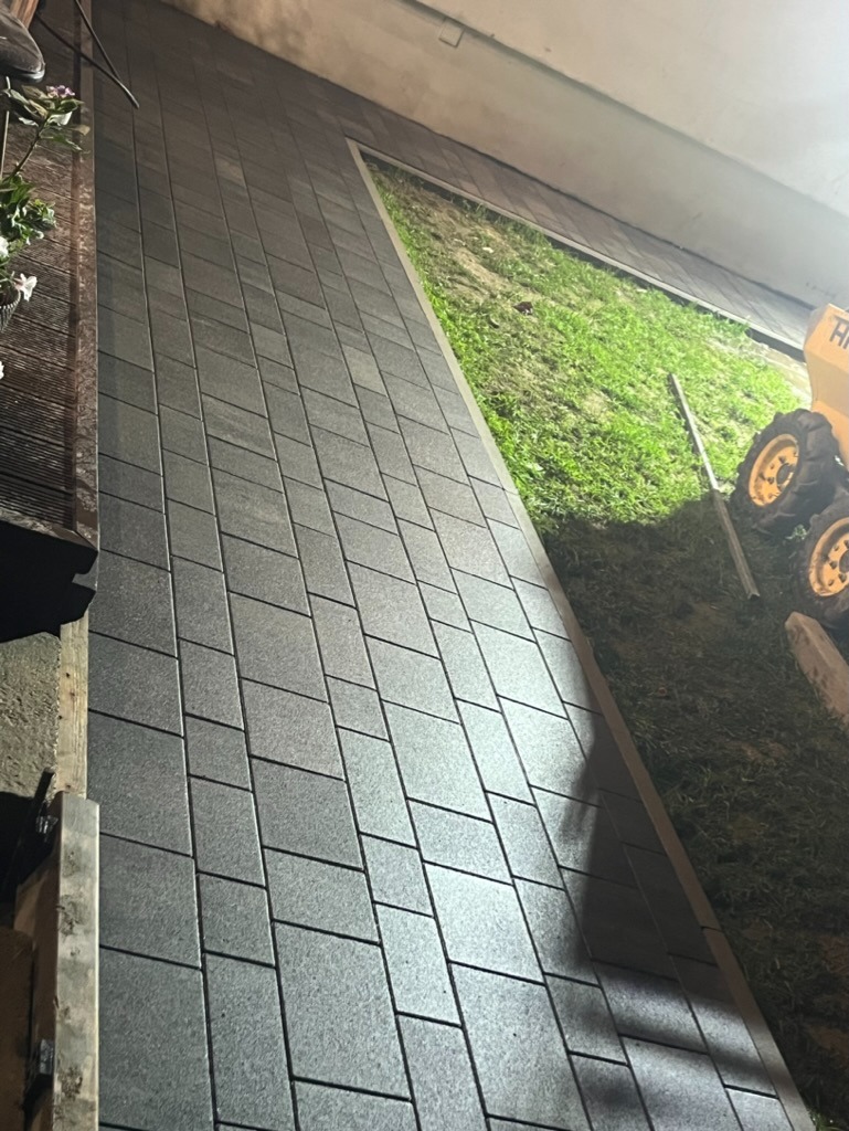 Ułożona szara kostka brukowa wzdłuż budynku z małym ogrodem, widoczny fragment maszyny budowlanej w tle.