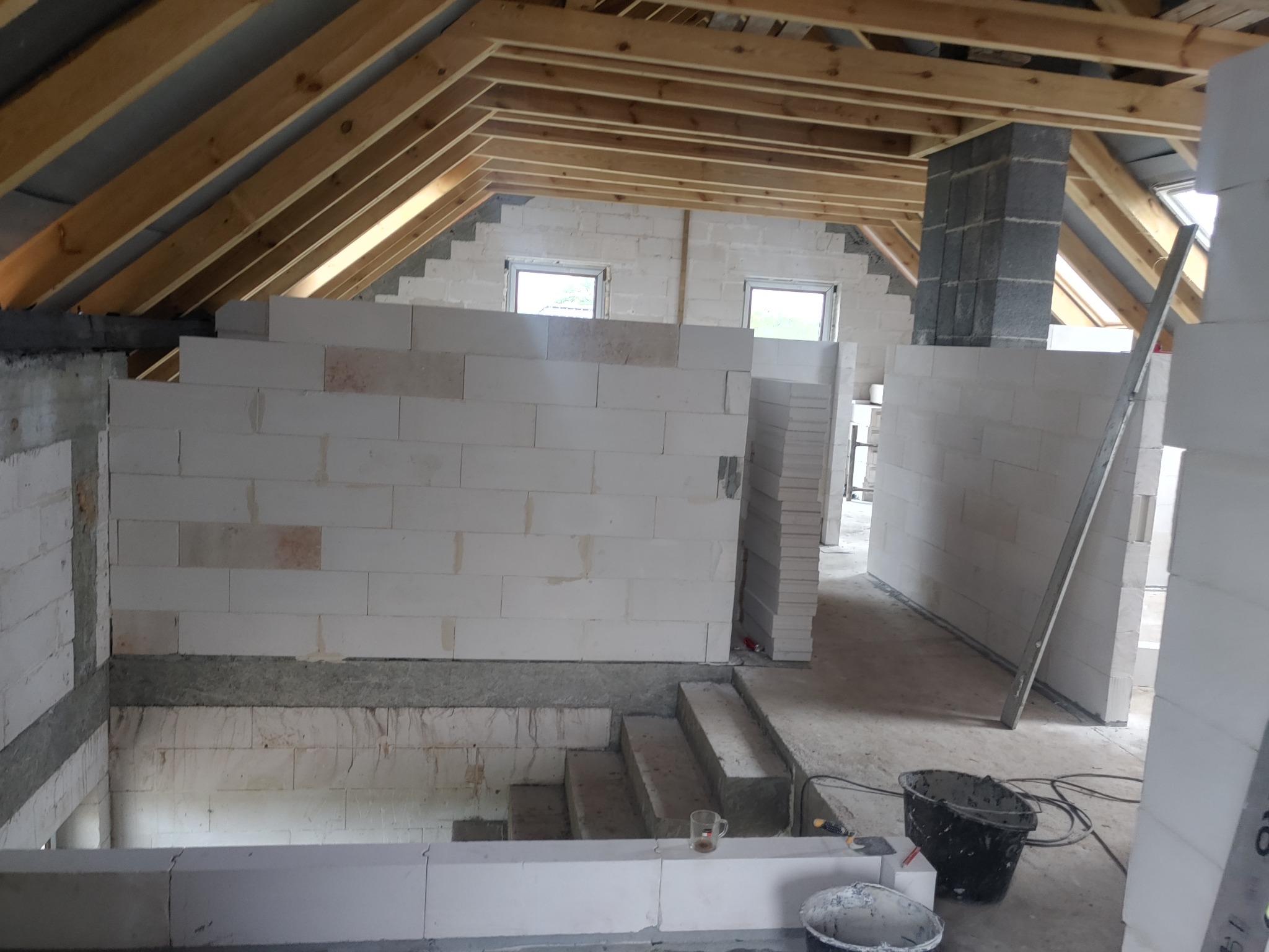 Wnętrze domu w trakcie budowy: wznoszone ściany z białych bloczków, widoczna drewniana konstrukcja dachu, okna oraz narzędzia budowlane.