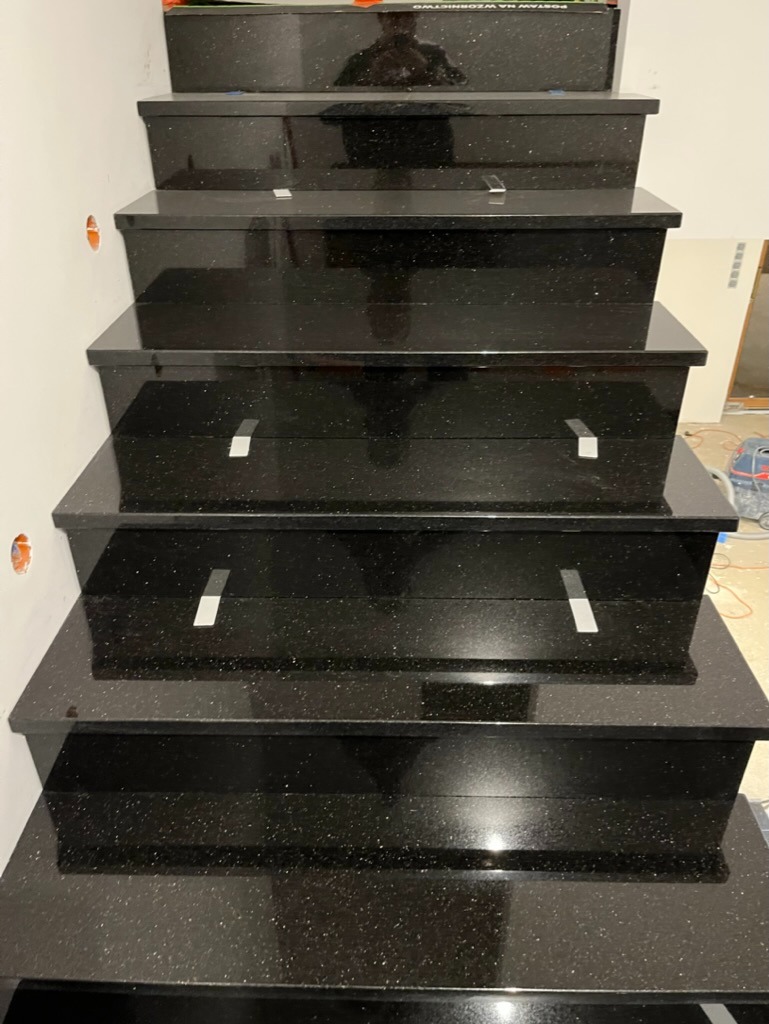 Schody wewnętrzne wykończone czarnym, błyszczącym kamieniem z drobnymi, jasnymi inkluzjami, widoczne srebrne listwy antypoślizgowe na krawędziach stopni, ściana z widocznymi puszkami elektrycznymi.