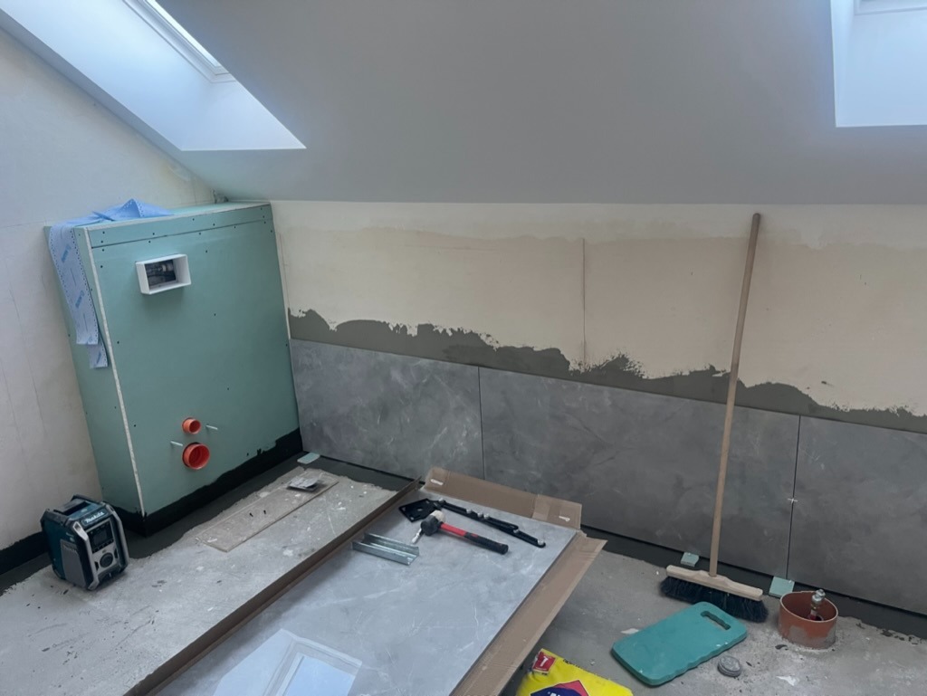 Wnętrze łazienki na poddaszu w trakcie prac wykończeniowych: zabudowa stelaża WC z płyt karton-gips, częściowo wyłożone szare płytki imitujące marmur, widoczne narzędzia i akcesoria budowlane, okno...