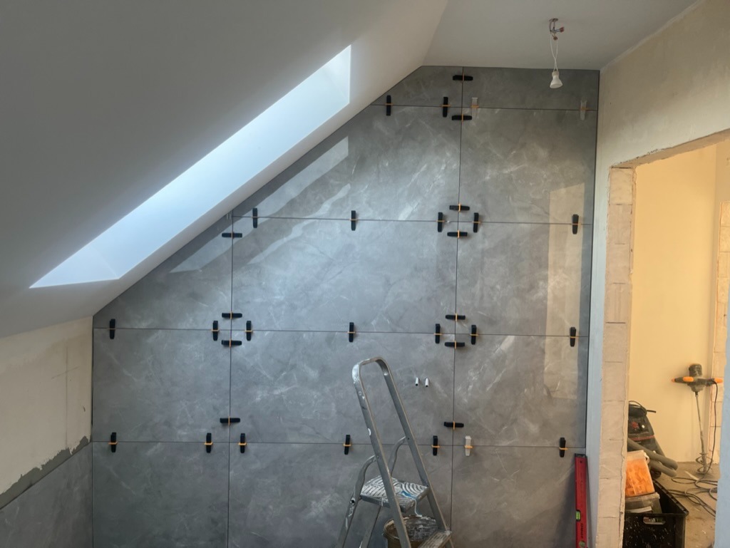 Montaż szarych płytek imitujących marmur na ścianie pod skosem poddasza w Choszcznie, widoczne krzyżyki dystansowe i drabina.