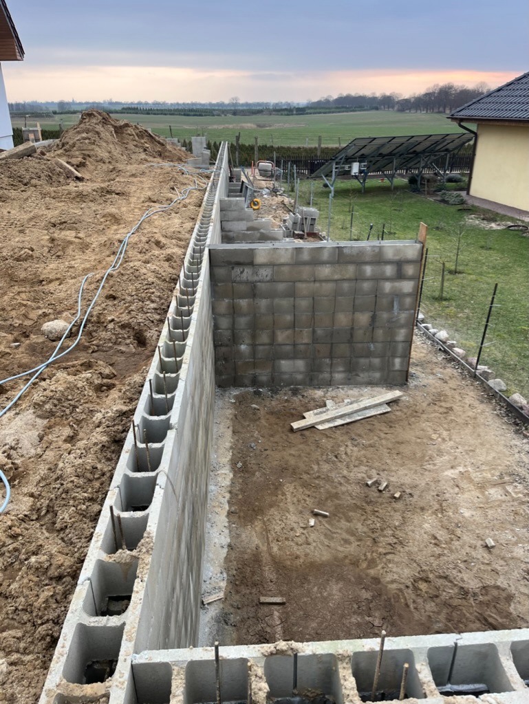 Wykop fundamentów z częściowo wzniesioną ścianą z bloczków betonowych, widoczne pręty zbrojeniowe, w tle hałda ziemi, panele słoneczne i budynek mieszkalny.