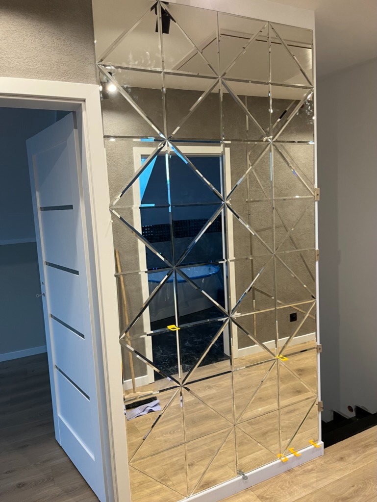 Ściana z dekoracyjnych luster w kształcie rombów, odbijająca wnętrze pokoju z widoczną białą ramą drzwi i fragmentem podłogi, zamontowana przy szarej ścianie.