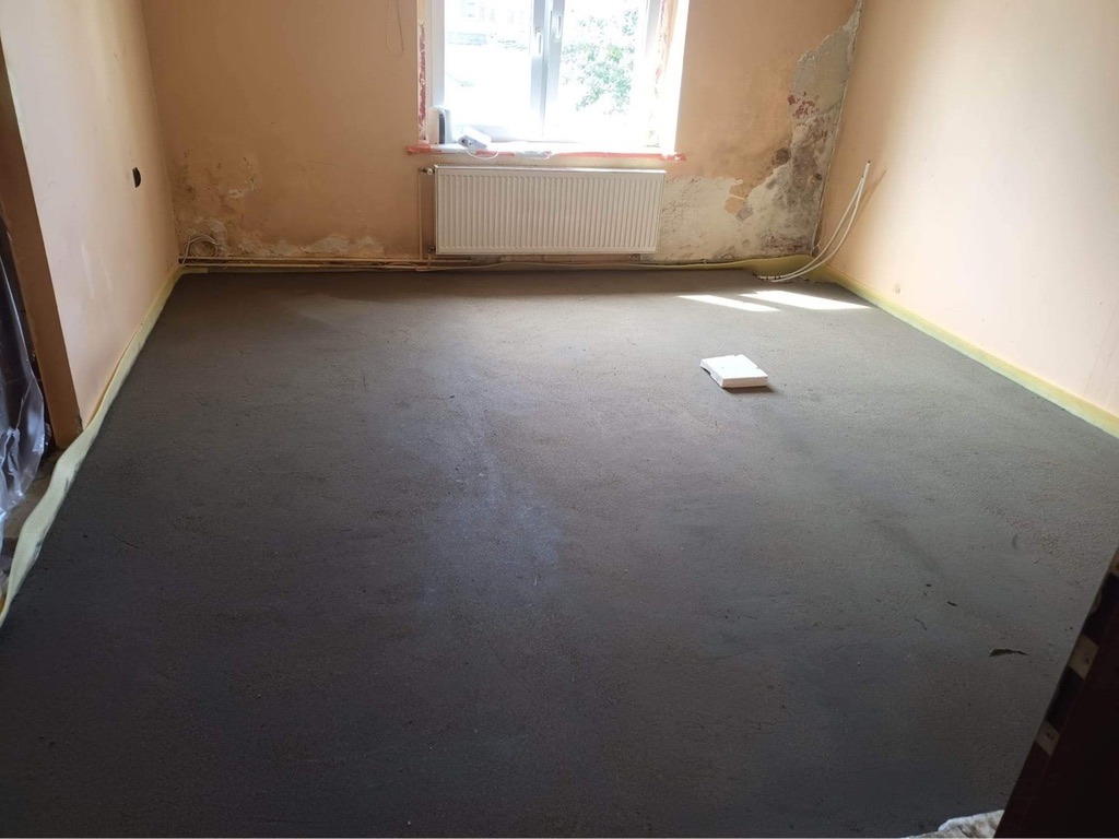 Świeżo wylana, ciemnoszara wylewka samopoziomująca w pokoju z przygotowanymi ścianami do dalszych prac wykończeniowych, widoczne zabezpieczenia folią przy ścianach i mały biały bloczek piankowy...