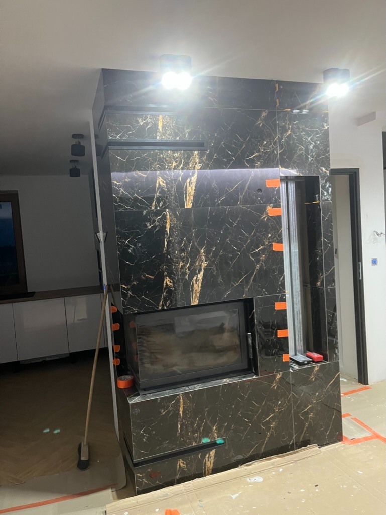 Montaż obudowy kominka z czarnego marmuru z złotymi żyłami, widoczne taśmy malarskie i częściowo zabezpieczona podłoga, oświetlenie punktowe sufitowe.