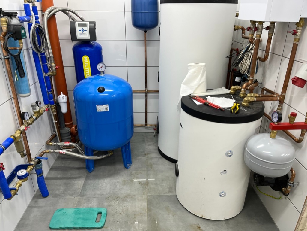 Węzeł hydroforowy z niebieskimi zbiornikami ciśnieniowymi i białymi zasobnikami ciepłej wody, widoczne rury miedziane i zawory, na jednym ze zbiorników leżą narzędzia: klucz nastawny i szczypce.