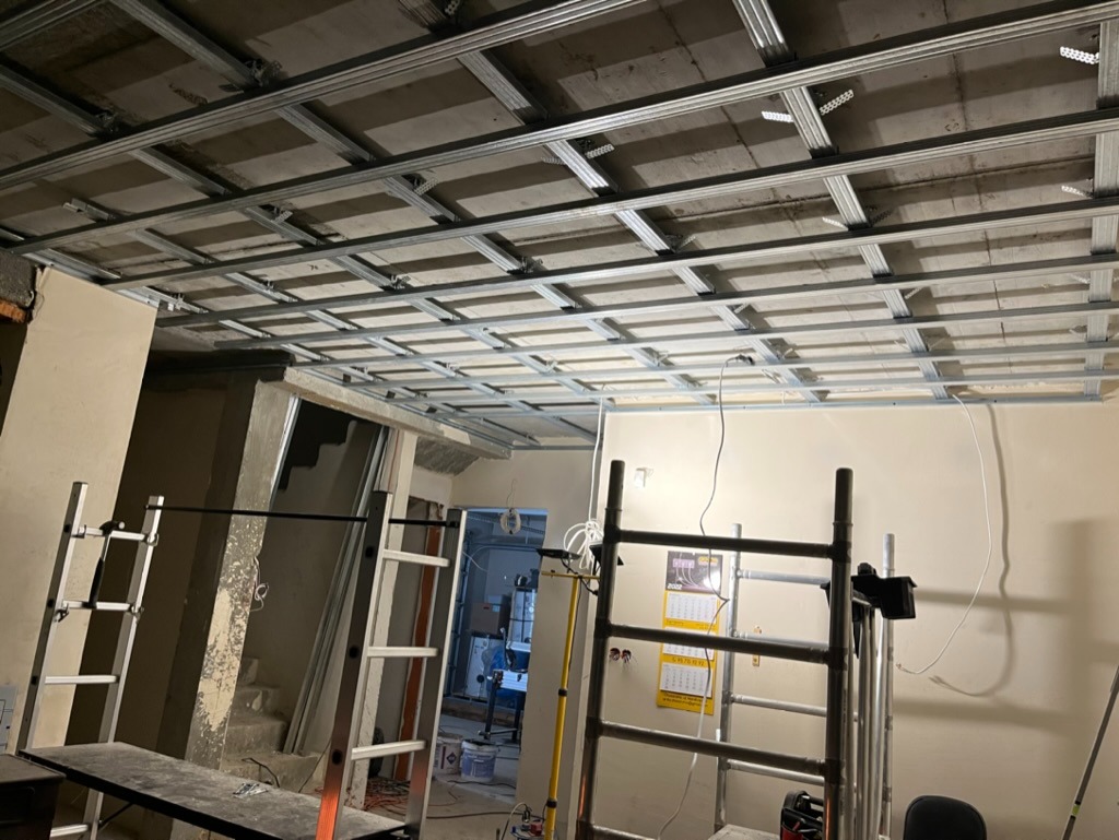 Metalowa konstrukcja sufitu podwieszanego w trakcie montażu, widoczne rusztowania i drabiny w remontowanym pomieszczeniu z widoczną instalacją elektryczną.