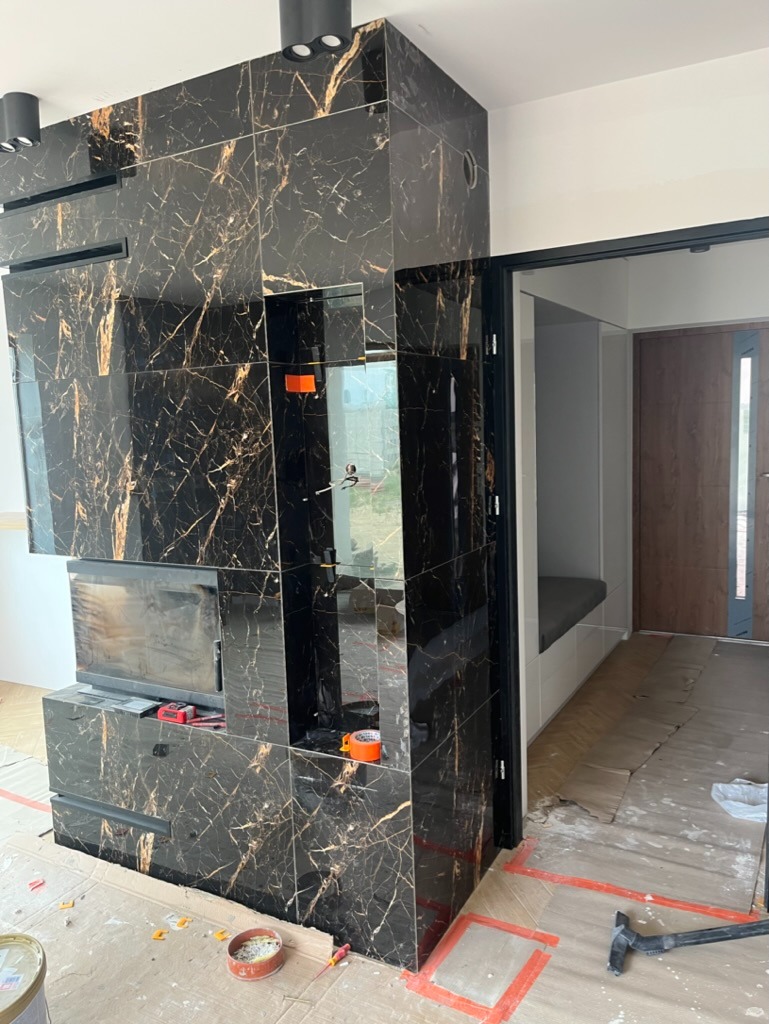 Obudowa kominka z czarnych, marmurkowych płyt ceramicznych w trakcie montażu, widoczne narzędzia i taśma malarska, w tle wejście do pomieszczenia z zabudowaną ławką.