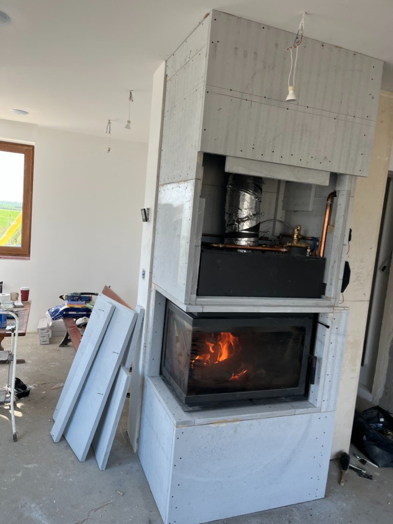 Wnętrze w trakcie wykończenia z zamontowanym, częściowo obudowanym kominkiem z palącym się ogniem, widoczne płyty kartonowo-gipsowe i elementy instalacji.