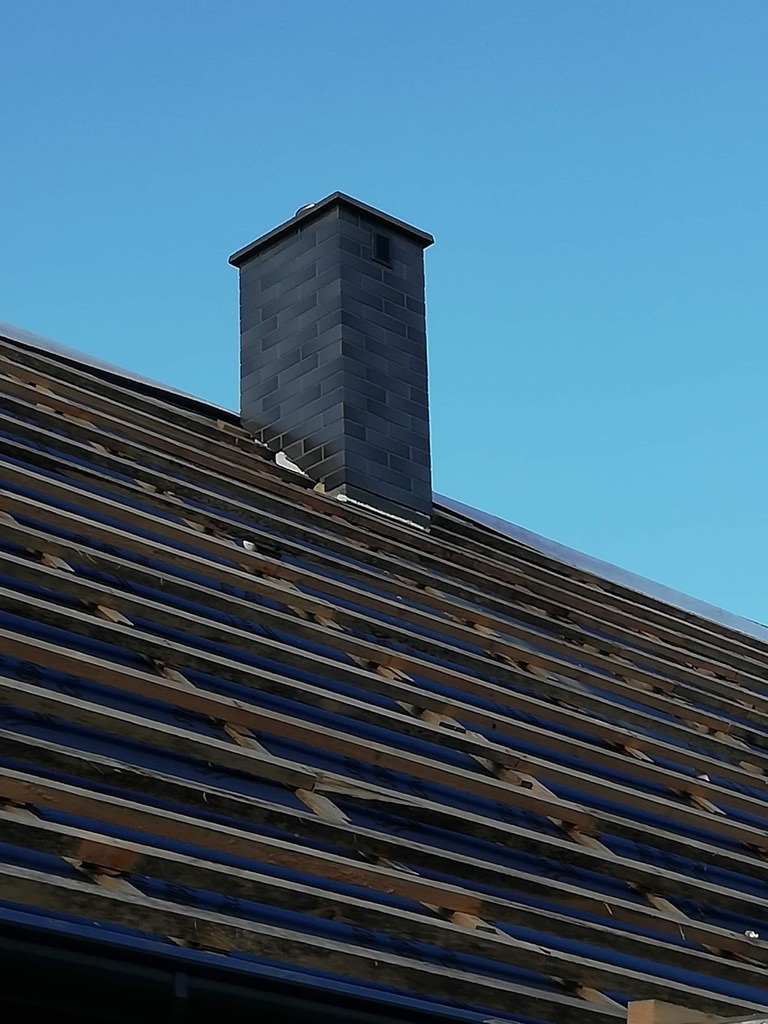 Ciemny, murowany komin na tle błękitnego nieba, widoczny na tle konstrukcji dachu z drewnianych łat pokrytych ciemną folią. Komin wykończony ciemnymi płytkami.