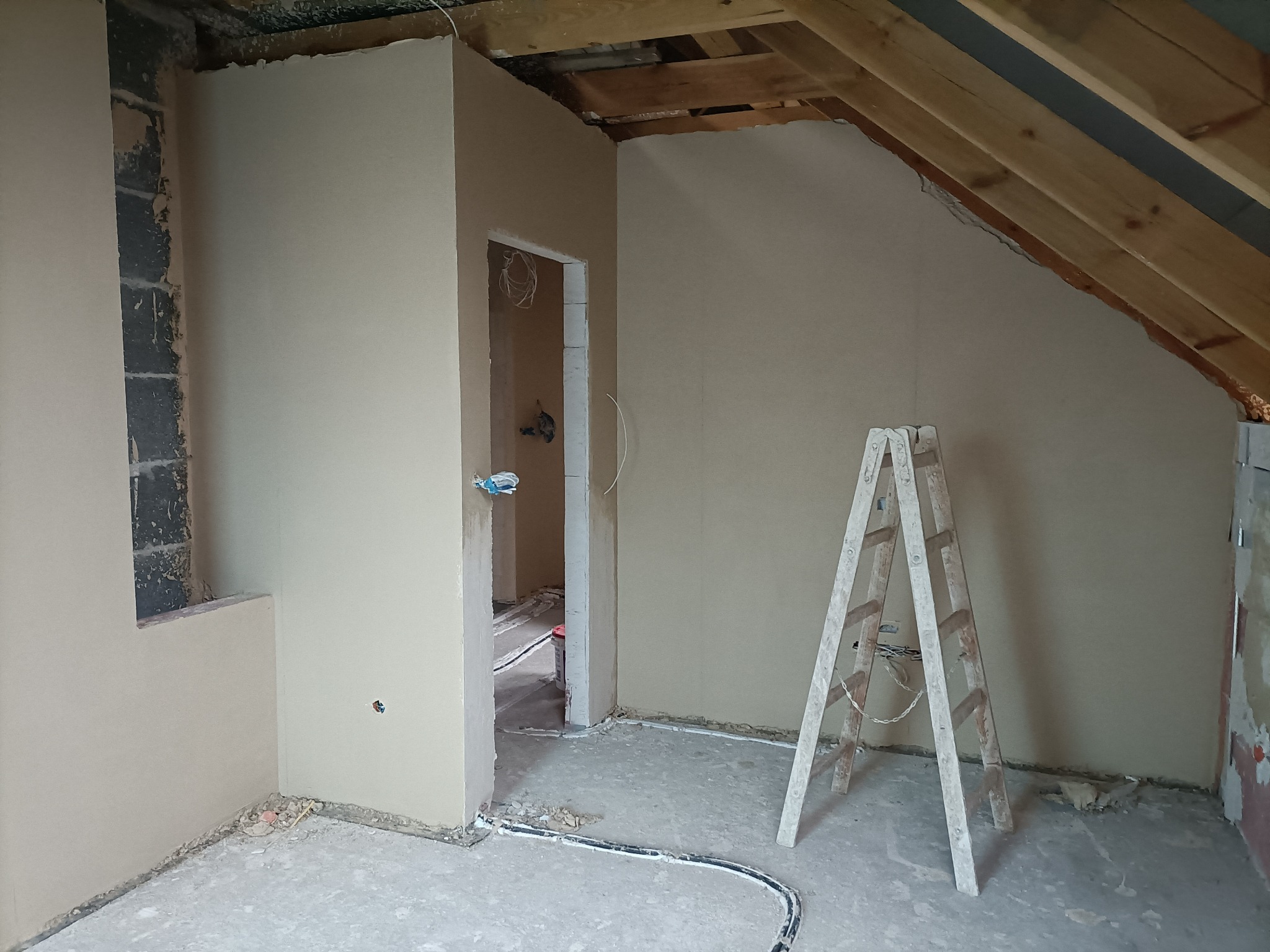 Poddasze w trakcie remontu: gładzie na ścianach, widoczne drewniane elementy konstrukcji dachu, drabina oparta o ścianę, instalacja elektryczna prowadzona po podłodze.