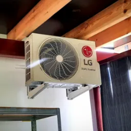Klimatyzator LG Dual Inverter zamontowany na ścianie w pomieszczeniu z drewnianą konstrukcją stropu, widoczne metalowe wsporniki mocujące i fragment regału.