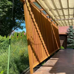 Pergola z drewnianą ścianą osłonową i rozwijanym materiałowym zadaszeniem, widok z boku na tle zieleni i fragmentu domu.