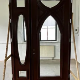 Ciemnobrązowe dwuskrzydłowe drzwi z szybami o nietypowych kształtach, opierające się o drewniane podpórki w surowym wnętrzu.