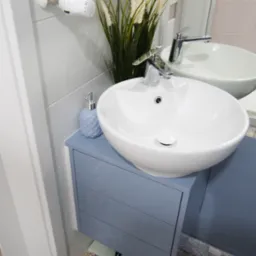 Łazienka z umywalką nablatową w kształcie misy, szafką w odcieniach szarości i wzorzystą podłogą z płytek ceramicznych; nad umywalką grzejnik drabinkowy i dekoracyjna trawa w wazonie.