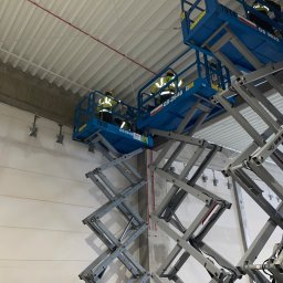 Oleksandr Konichenko - Dwóch elektryków w żółtych kamizelkach na podnośnikach koszowych montuje instalacje elektryczne pod sufitem w hali przemysłowej.