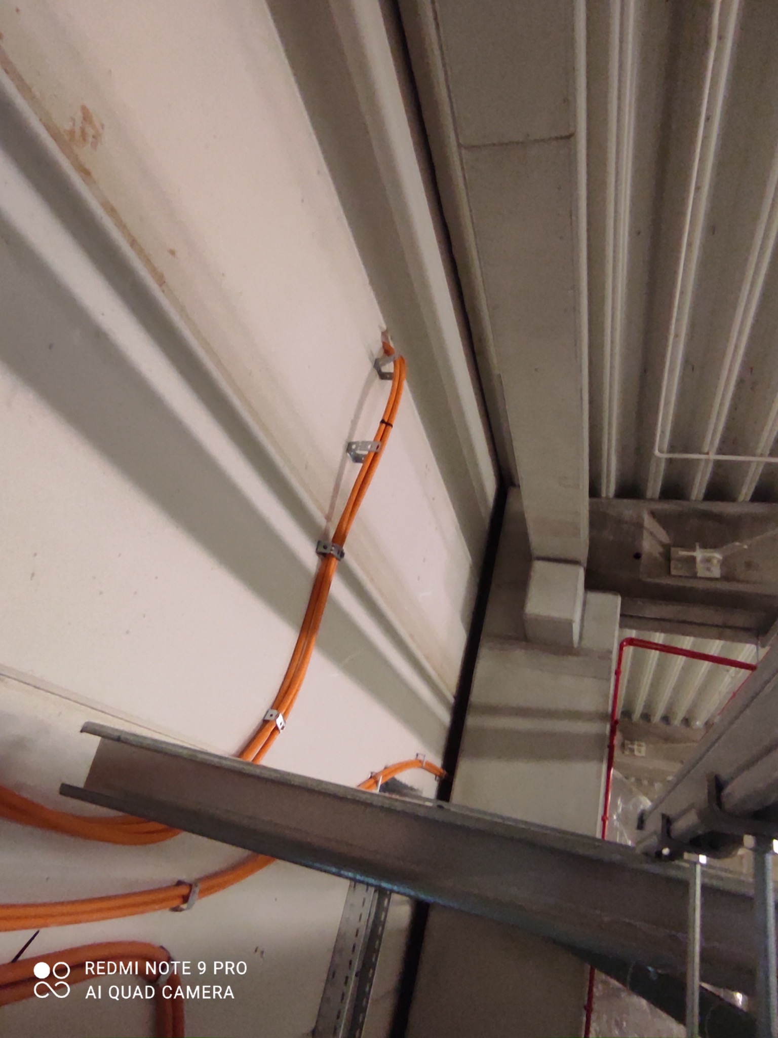 Pomarańczowe przewody elektryczne zamocowane do ściany za pomocą metalowych uchwytów, widoczne szyny montażowe pod sufitem w hali przemysłowej.