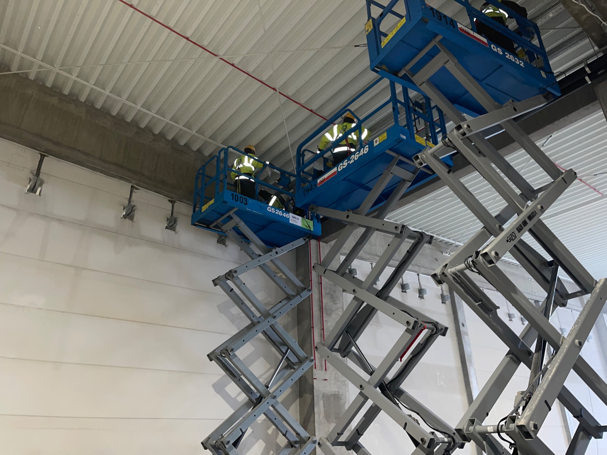 Dwóch elektryków w żółtych kamizelkach na podnośnikach koszowych montuje instalacje elektryczne pod sufitem w hali przemysłowej.