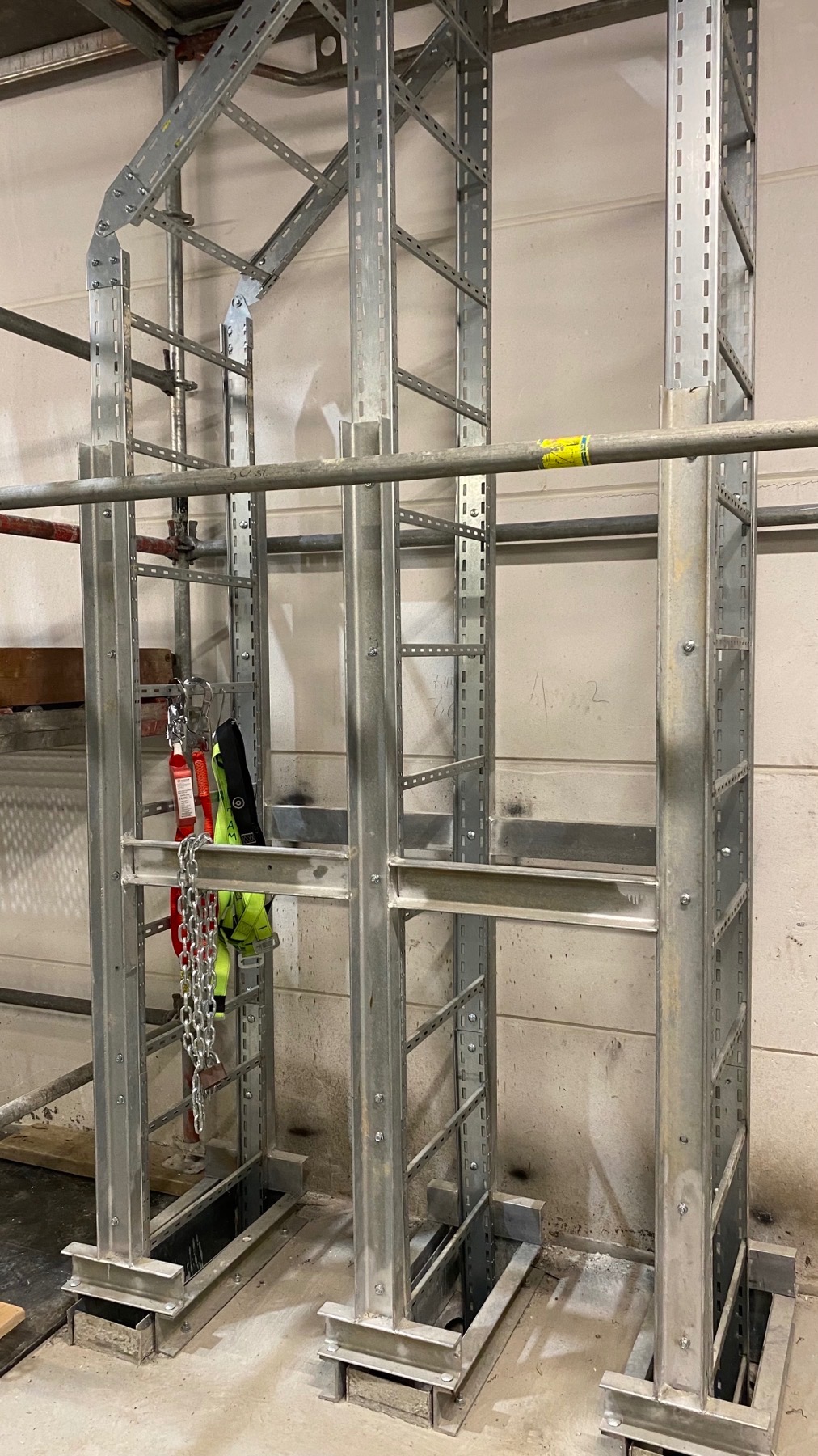 Metalowa konstrukcja wsporcza z perforowanymi profilami, prawdopodobnie pod kable lub rury, zamontowana w surowym wnętrzu. Na konstrukcji zawieszony sprzęt roboczy: uprząż asekuracyjna i łańcuch.