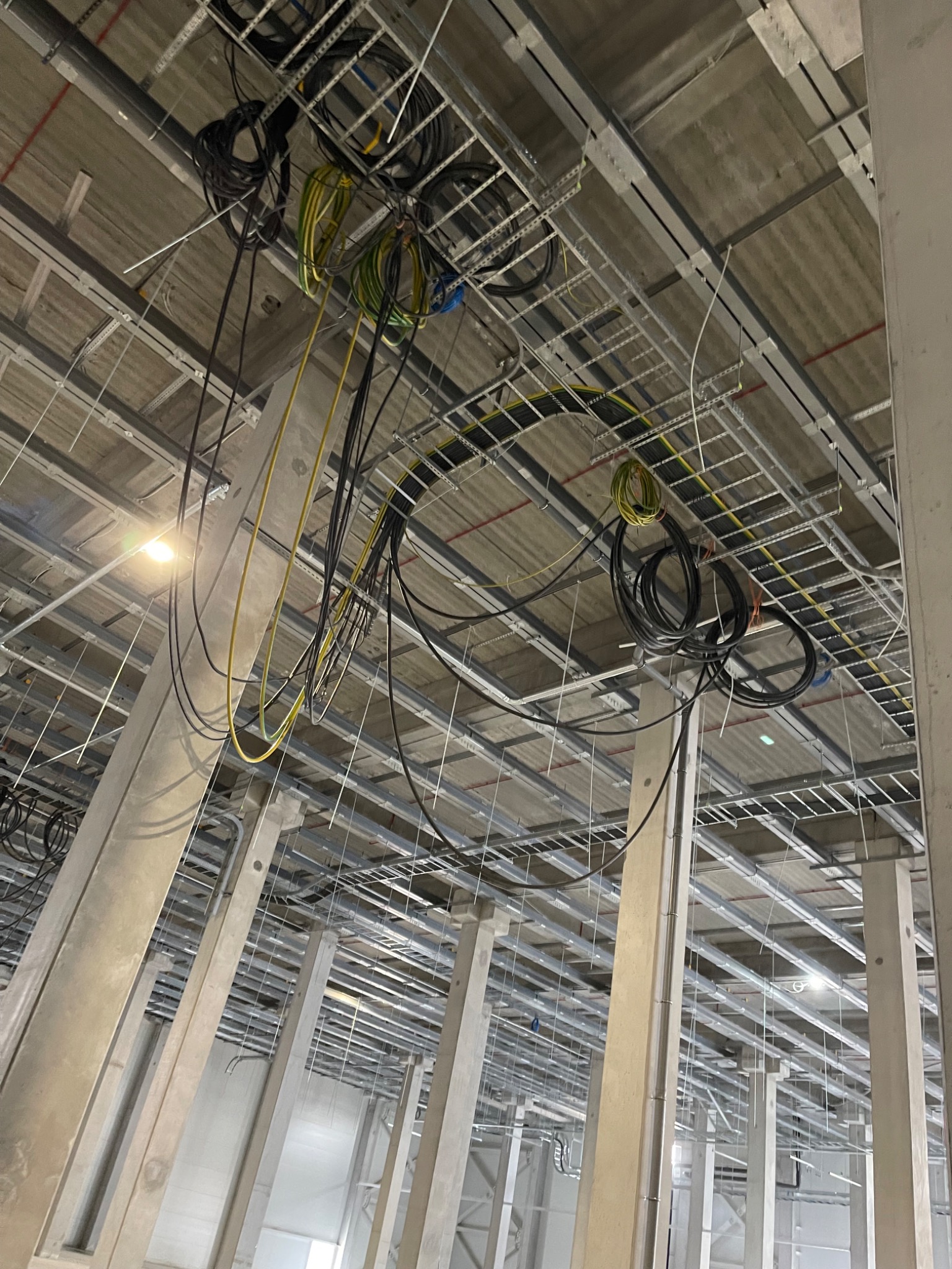 Kable elektryczne zwisające z metalowych korytek pod sufitem w hali przemysłowej, widoczne betonowe kolumny i oświetlenie.