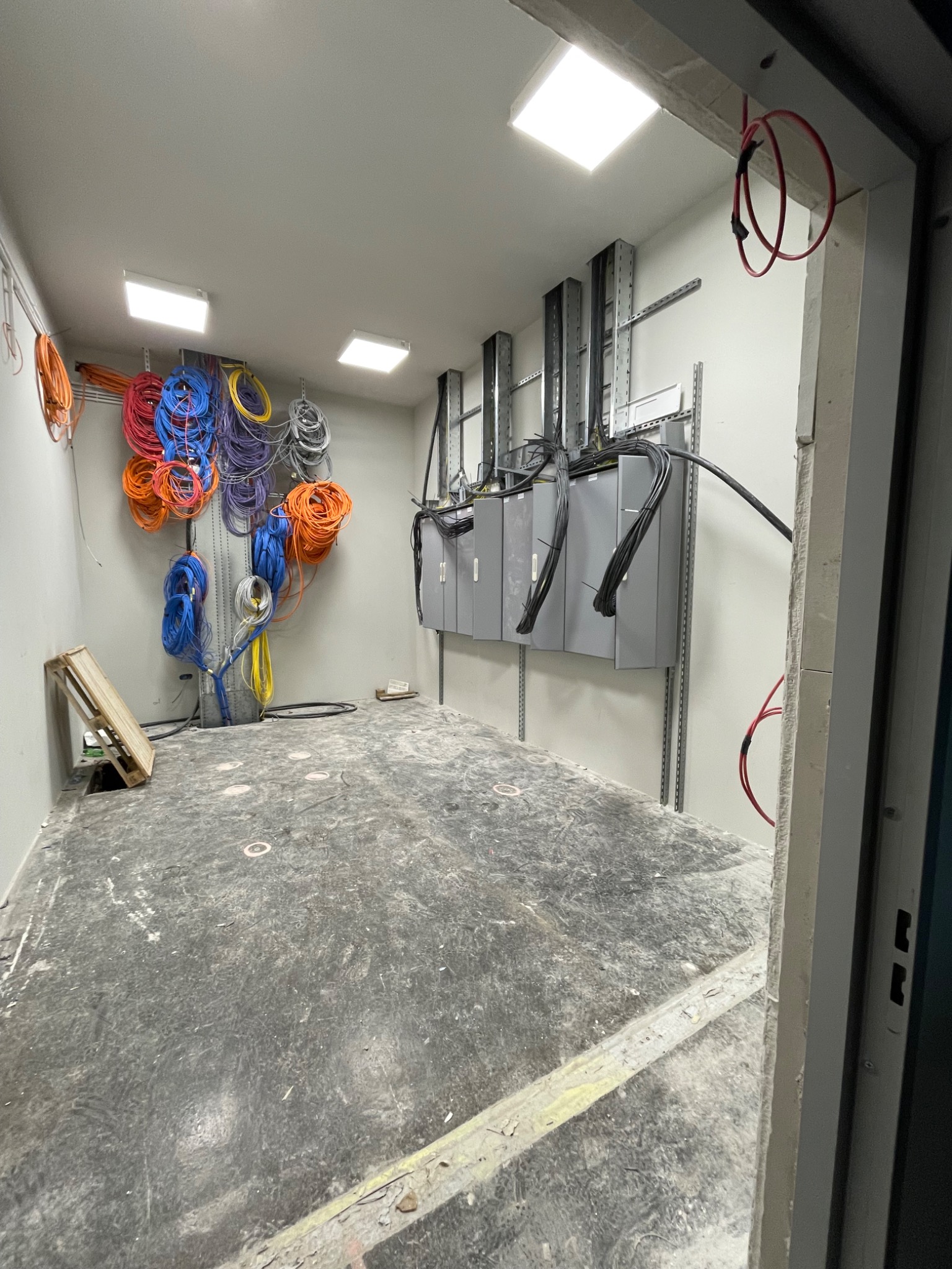 Wnętrze pomieszczenia w trakcie instalacji elektrycznej: ściany z zamontowanymi skrzynkami elektrycznymi i metalowymi szynami, zwisające kable różnej grubości i koloru, surowa podłoga, paleta...