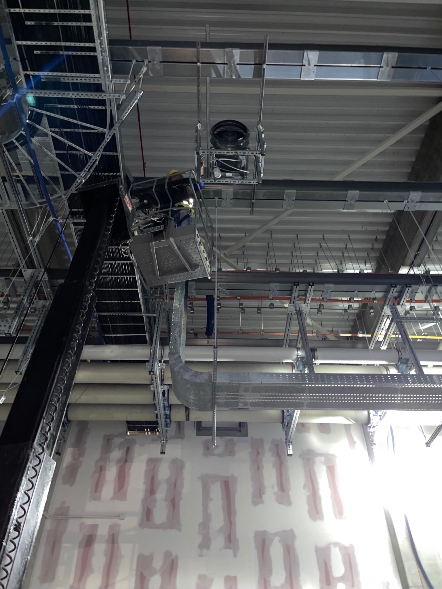 Pracownik w podnośniku koszowym instaluje system wentylacyjny pod sufitem hali z widocznymi elementami konstrukcyjnymi i instalacjami.