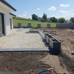 HubMar - Teren wokół domu w trakcie prac ziemnych i brukarskich: przygotowanie podłoża, rozsypany żwir, ustawione donice, widoczne fundamenty i ułożony fragment obrzeża z kostki granitowej.