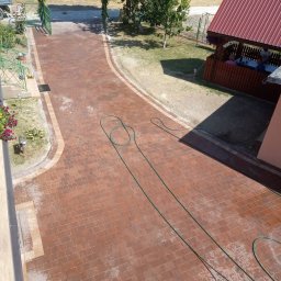 HubMar - Widok z góry na nowo ułożoną, czerwoną kostkę brukową na podjeździe, z zielonym wężem ogrodowym leżącym na powierzchni. Z tyłu altana z ludźmi i brama z zieleni. Widoczne ślady mycia kostki.