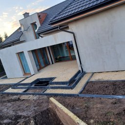 HubMar - Taras domu jednorodzinnego z wysypanym żwirem, obrzeża z betonowych kostek, przygotowane rabaty ziemne, widok na elewację budynku z oknami.