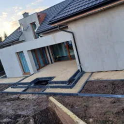 Taras domu jednorodzinnego z wysypanym żwirem, obrzeża z betonowych kostek, przygotowane rabaty ziemne, widok na elewację budynku z oknami.