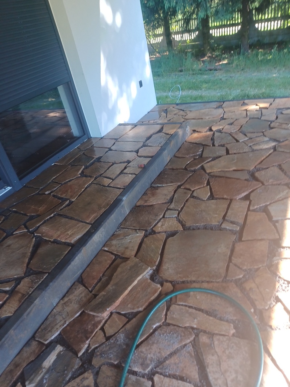 Ułożona z nieregularnych, brązowo-złotych płyt kamiennych nawierzchnia tarasu przy budynku z szarą stolarką okienną i roletą, widoczny zielony wąż ogrodowy.
