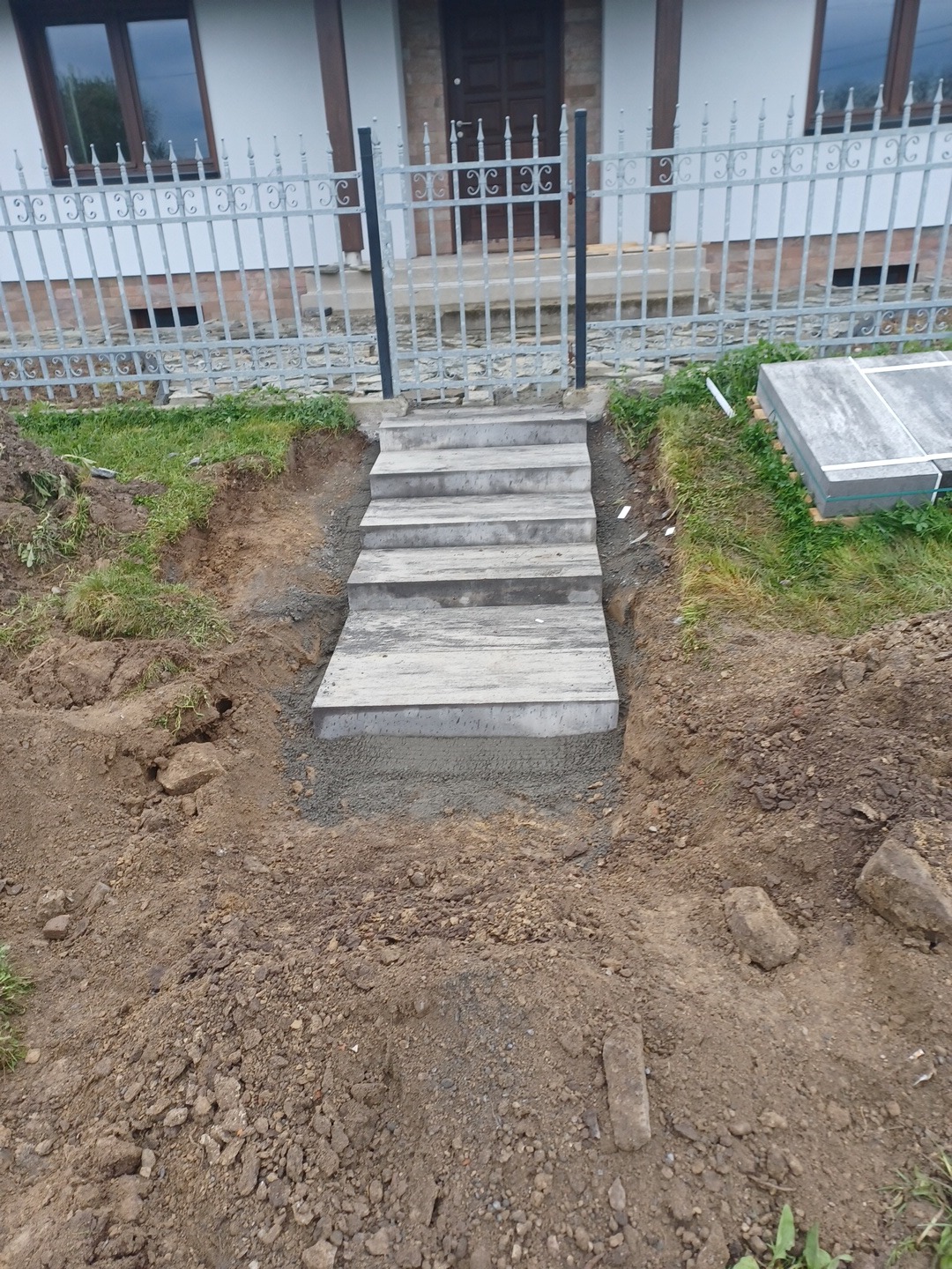 Nowe betonowe schody prowadzące do domu, widoczne ślady prac ziemnych i fragment ogrodzenia.