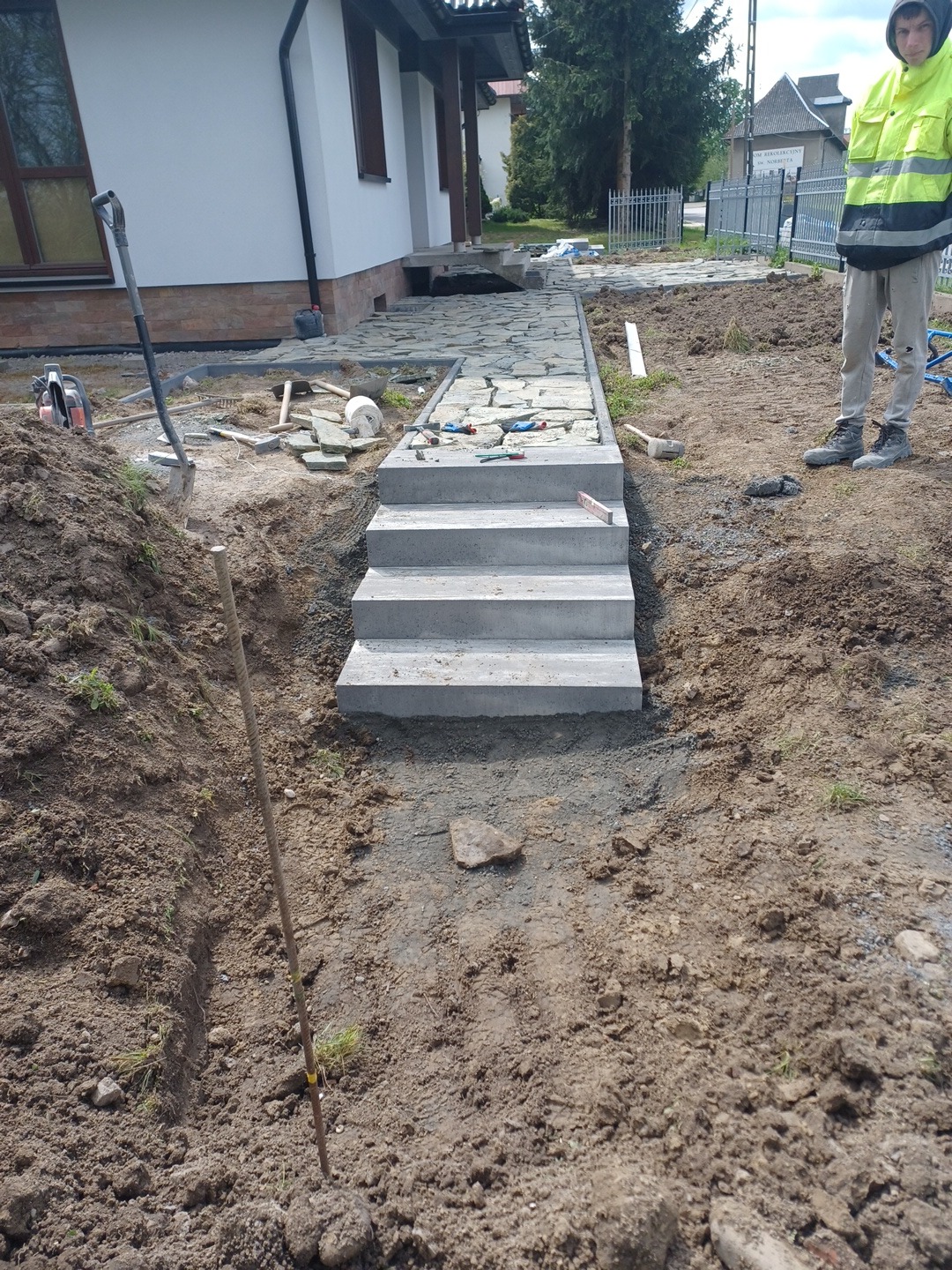 Budowa ścieżki z kamienia i betonowych schodów przed domem jednorodzinnym, widoczny pracownik w odblaskowej kurtce i narzędzia brukarskie.