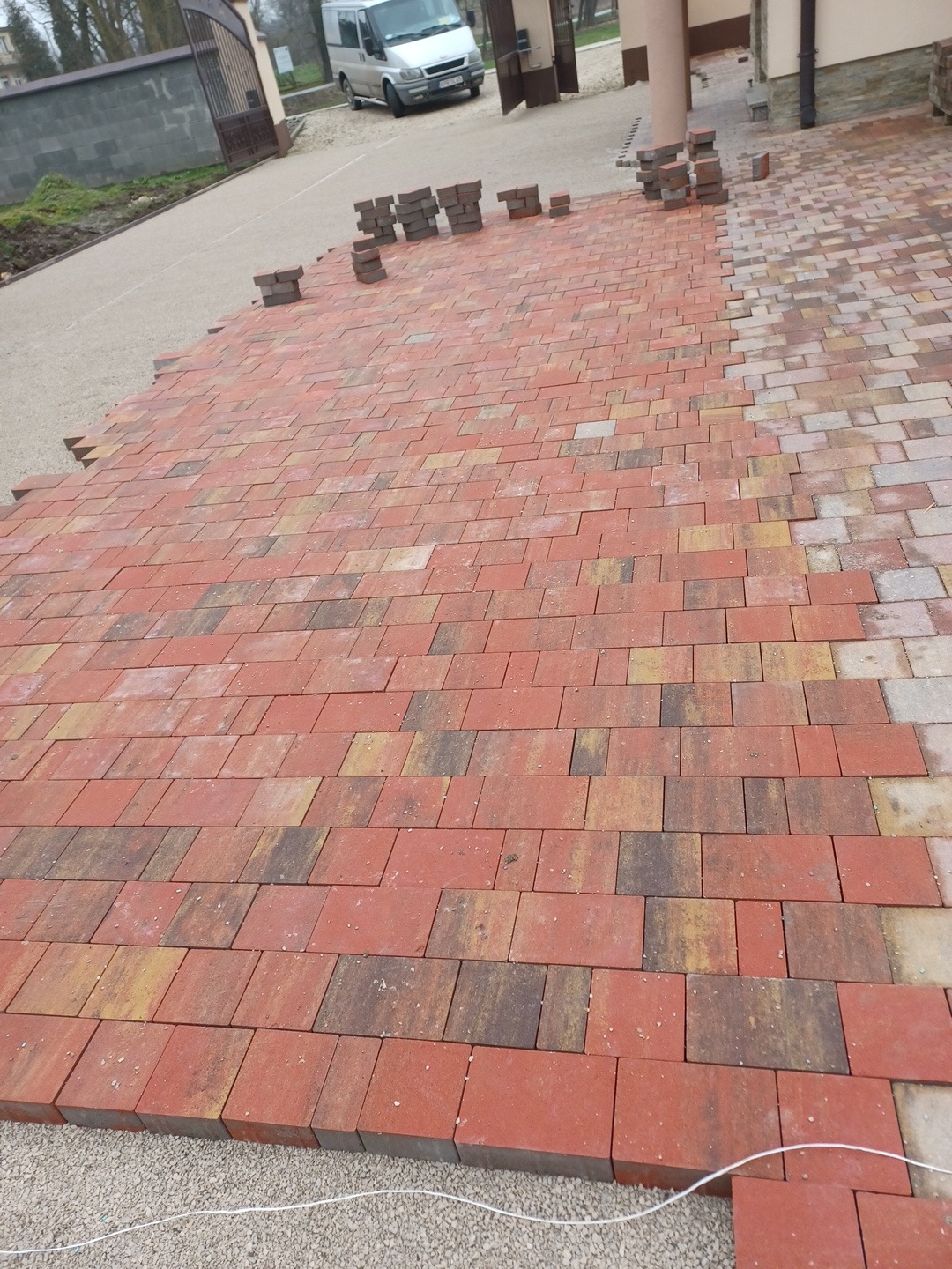 Układanie czerwono-brązowej kostki brukowej na podsypce piaskowej, widoczny fragment ułożonej powierzchni oraz stosy kostki przygotowanej do dalszych prac, biały sznurek wyznaczający linię układania.