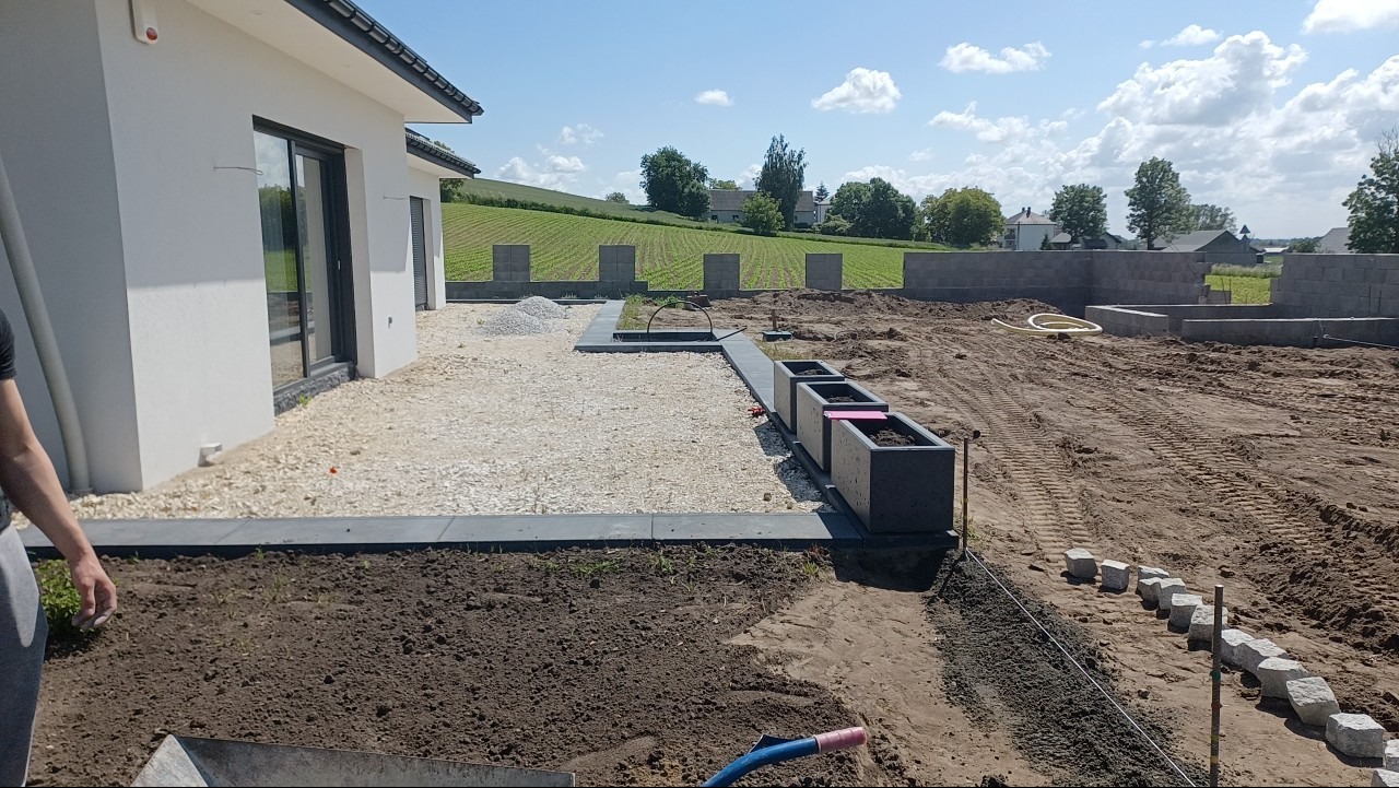 Teren wokół domu w trakcie prac ziemnych i brukarskich: przygotowanie podłoża, rozsypany żwir, ustawione donice, widoczne fundamenty i ułożony fragment obrzeża z kostki granitowej.